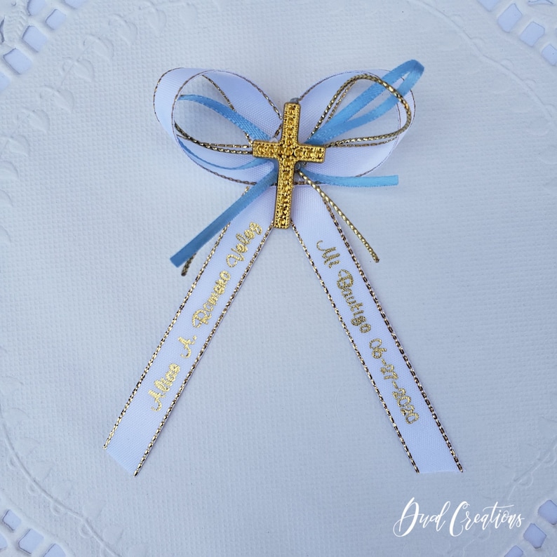 May include: A decorative bow with a gold cross centerpiece. The bow is white with light blue accents and gold trim. The ribbons have gold text that reads "Alice A. Domingo Velez" and "Mi Bautizo 06-27-2020".