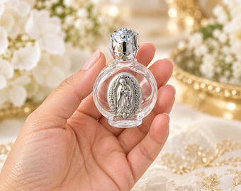 Puede incluir: Un frasco pequeño de vidrio transparente con cuerpo redondo y tapón plateado en forma de rosa. La parte frontal del frasco presenta un medallón ovalado de metal plateado que representa a la Virgen de Guadalupe en relieve sobre el vidrio.