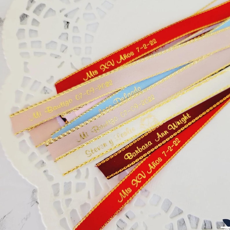 May include: Assorted satin ribbons in red, pink, blue, and burgundy, with gold trim and personalized text. The ribbons feature phrases like "Mis XV A&ntilde;os" and names with dates, ideal for event favors or decorations.