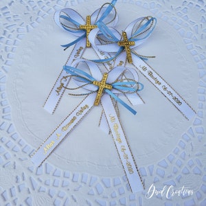 May include: Three baptism favors with white and light blue ribbons, gold crosses, and gold text. The text on the ribbons reads "Alias A. Romero Velaz" and "Mi Bautizo 06-27-2020".