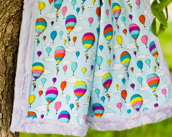 May include: A light blue baby blanket featuring a colorful hot air balloon pattern. The balloons are in various colors, including yellow, orange, pink, and blue. The blanket has a soft, lavender-colored border.