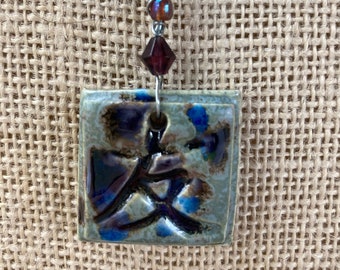 May include: A silver chain necklace with a square ceramic pendant featuring a blue and brown abstract design. The pendant is attached to the chain with a dark red crystal and a brown iridescent bead.