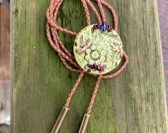 May include: A bolo tie featuring a round, light green ceramic pendant with an embossed floral pattern. The pendant is accented with small, iridescent purple and blue beads. It hangs from a braided brown leather cord with two metallic end tips.