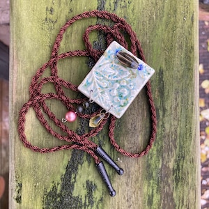 Könnte beinhalten: Eine handgefertigte Bolo-Krawatte mit einem quadratischen, cremefarbenen Keramikanhänger mit einem blau-grünen Blumenmuster. Der Anhänger ist mit einer braunen und einer blauen Perle verziert. Die Bolo-Krawatte hat eine braune Kordel und schwarze Metallspitzen.