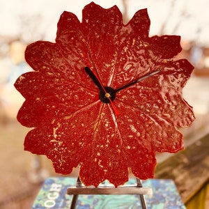 May include: A red leaf-shaped clock with gold accents on a metal easel. The clock hands are black and gold.