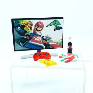 May include: A miniature gaming setup with a flat-screen monitor showing a racing game with Mario. A red controller, a yellow snack bag, a bottle of soda, and a candy cane are on a clear acrylic table. The background is white.