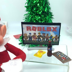 May include: A miniature scene with a television showing "ROBLOX" characters. A bag of crisps, a bottle of fizzy drink, and a chocolate bar are on a clear table. A green tinsel Christmas tree is in the background.