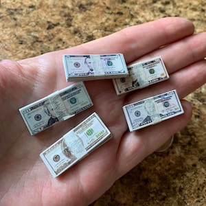 Może przedstawiać: Pięć stos&oacute;w miniaturowych banknot&oacute;w dolar&oacute;w amerykańskich, w tym nominały 5, 10, 20, 50 i 100 dolar&oacute;w, jest wyświetlanych na dłoni. Banknoty są nadrukowane wizerunkiem Abrahama Lincolna, Alexandra Hamiltona, Andrew Jacksona, Ulyssesa S. Granta i Benjamina Franklina.