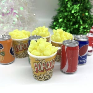 May include: Miniature popcorn buckets filled with yellow popcorn, alongside soda cans of Fanta, Coca-Cola, and Pepsi. The popcorn buckets have a classic design. The scene is festive, with a Christmas tree in the background.