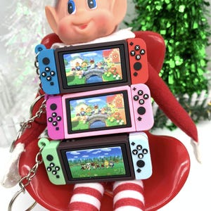 May include: Three miniature Nintendo Switch keychains stacked on top of each other. The top keychain is red, the middle keychain is pink, and the bottom keychain is green. Each keychain has a different screen image of a video game.