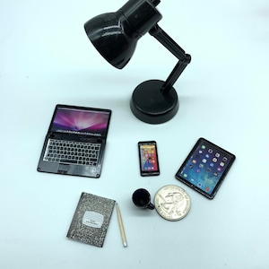 Puede incluir: Una l&aacute;mpara de escritorio negra con una pantalla negra, una computadora port&aacute;til negra, una tableta negra, un tel&eacute;fono inteligente negro, una taza de caf&eacute; negra, un l&aacute;piz y una moneda de plata sobre una superficie blanca.