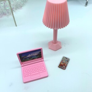 Puede incluir: Un ordenador port&aacute;til de juguete rosa, un tel&eacute;fono m&oacute;vil de juguete rosa y una l&aacute;mpara de juguete rosa con una pantalla plisada. El port&aacute;til tiene una pantalla que muestra una escena tropical.