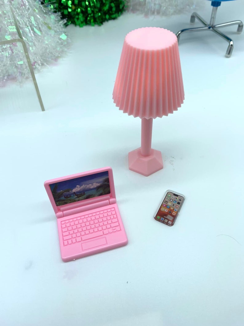 Puede incluir: Un ordenador port&aacute;til de juguete rosa, un tel&eacute;fono m&oacute;vil de juguete rosa y una l&aacute;mpara de juguete rosa con una pantalla plisada. El port&aacute;til tiene una pantalla que muestra una escena tropical.