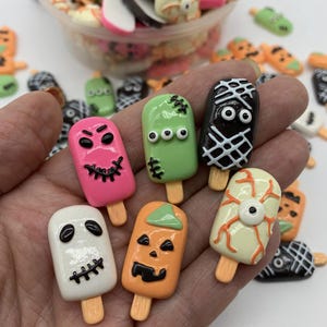 May include: A collection of Halloween-themed popsicle-shaped charms in various colors. The charms feature designs like a mummy, a zombie, a pumpkin, and a skull. Some charms are pink, green, orange, white, and black. The charms are on wooden sticks.