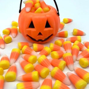Puede incluir: Un cubo de calabaza naranja de pl&aacute;stico lleno de caramelos de ma&iacute;z. El cubo tiene un asa negra y una cara tallada. Los caramelos de ma&iacute;z est&aacute;n esparcidos alrededor del cubo. Los caramelos son amarillos, naranjas y blancos.