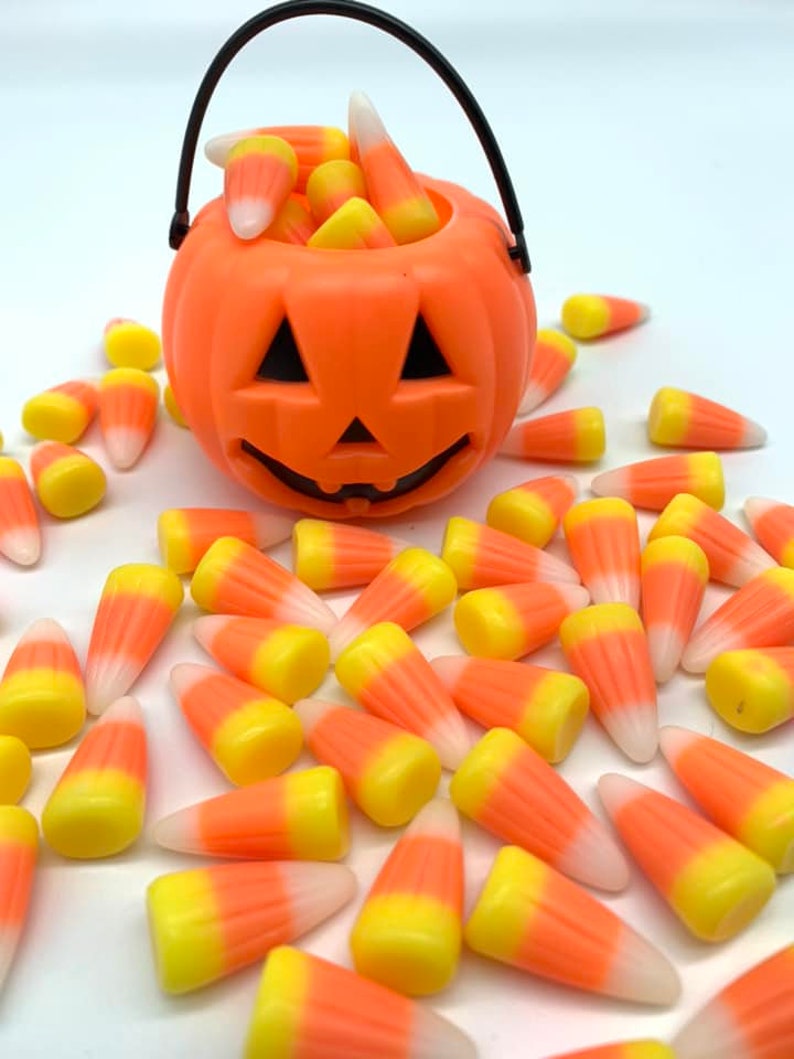 Puede incluir: Un cubo de calabaza naranja de pl&aacute;stico lleno de caramelos de ma&iacute;z. El cubo tiene un asa negra y una cara tallada. Los caramelos de ma&iacute;z est&aacute;n esparcidos alrededor del cubo. Los caramelos son amarillos, naranjas y blancos.