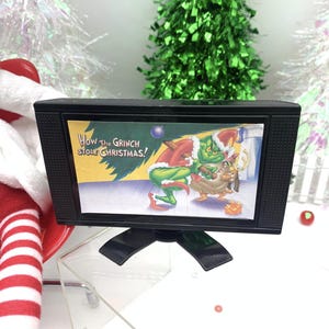 May include: A small black television set featuring a scene from "How the Grinch Stole Christmas!". The TV is on a stand, set against a festive backdrop with Christmas trees and decorations, evoking a holiday theme.