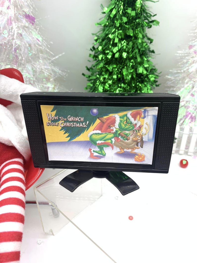 May include: A small black television set featuring a scene from "How the Grinch Stole Christmas!". The TV is on a stand, set against a festive backdrop with Christmas trees and decorations, evoking a holiday theme.