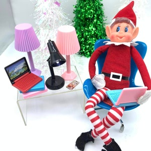 Puede incluir: Un mu&ntilde;eco de duende de Navidad sentado en una silla azul con un ordenador port&aacute;til rosa. El duende lleva un atuendo a rayas rojas y blancas y zapatos negros. Hay dos l&aacute;mparas rosas y una l&aacute;mpara de escritorio negra sobre una mesa de cristal transparente con un ordenador port&aacute;til rojo y rosa y un tel&eacute;fono rosa.