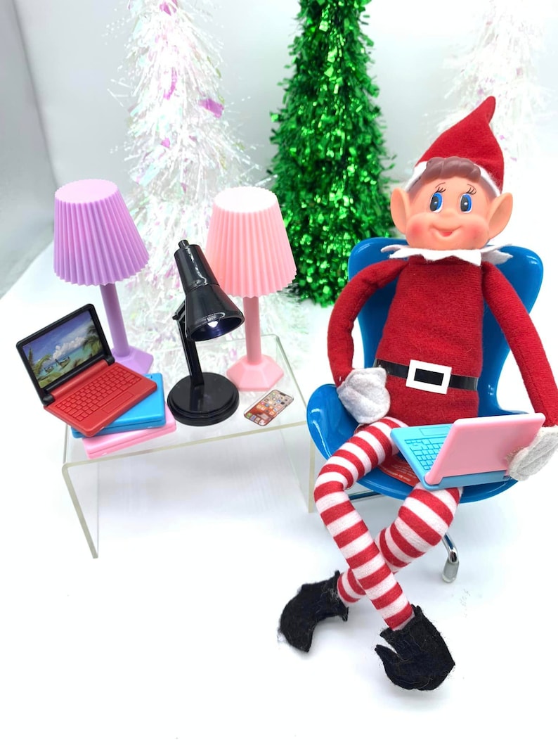 Puede incluir: Un mu&ntilde;eco de duende de Navidad sentado en una silla azul con un ordenador port&aacute;til rosa. El duende lleva un atuendo a rayas rojas y blancas y zapatos negros. Hay dos l&aacute;mparas rosas y una l&aacute;mpara de escritorio negra sobre una mesa de cristal transparente con un ordenador port&aacute;til rojo y rosa y un tel&eacute;fono rosa.