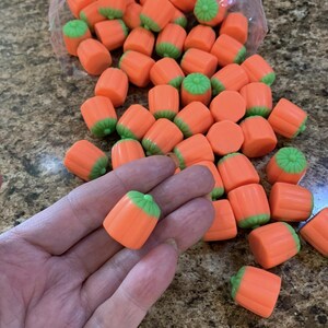 Puede incluir: Caramelos con forma de calabaza, de color naranja y verde. Los caramelos son cil&iacute;ndricos, con cuerpos naranjas y tapas verdes, que se asemejan a calabazas en miniatura. Se ve un pu&ntilde;ado de caramelos, y m&aacute;s se derraman de una bolsa transparente.
