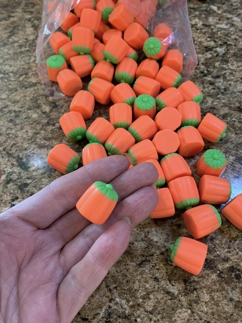Puede incluir: Caramelos con forma de calabaza, de color naranja y verde. Los caramelos son cil&iacute;ndricos, con cuerpos naranjas y tapas verdes, que se asemejan a calabazas en miniatura. Se ve un pu&ntilde;ado de caramelos, y m&aacute;s se derraman de una bolsa transparente.