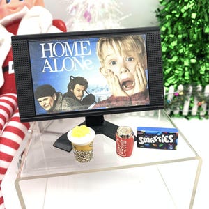 May include: Miniature black television showing the film "Home Alone", alongside a small soda can, popcorn, and a box of Smarties sweets. The items are on a clear acrylic table, with a Christmas tree and elf in the background.