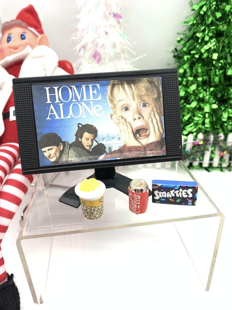 May include: Miniature black television showing the film "Home Alone", alongside a small soda can, popcorn, and a box of Smarties sweets. The items are on a clear acrylic table, with a Christmas tree and elf in the background.