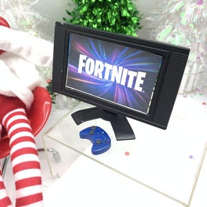 May include: Miniature black television displaying the word "FORTNITE". A blue game controller is positioned below the screen. The TV and controller are on a clear acrylic surface, with Christmas trees in the background.
