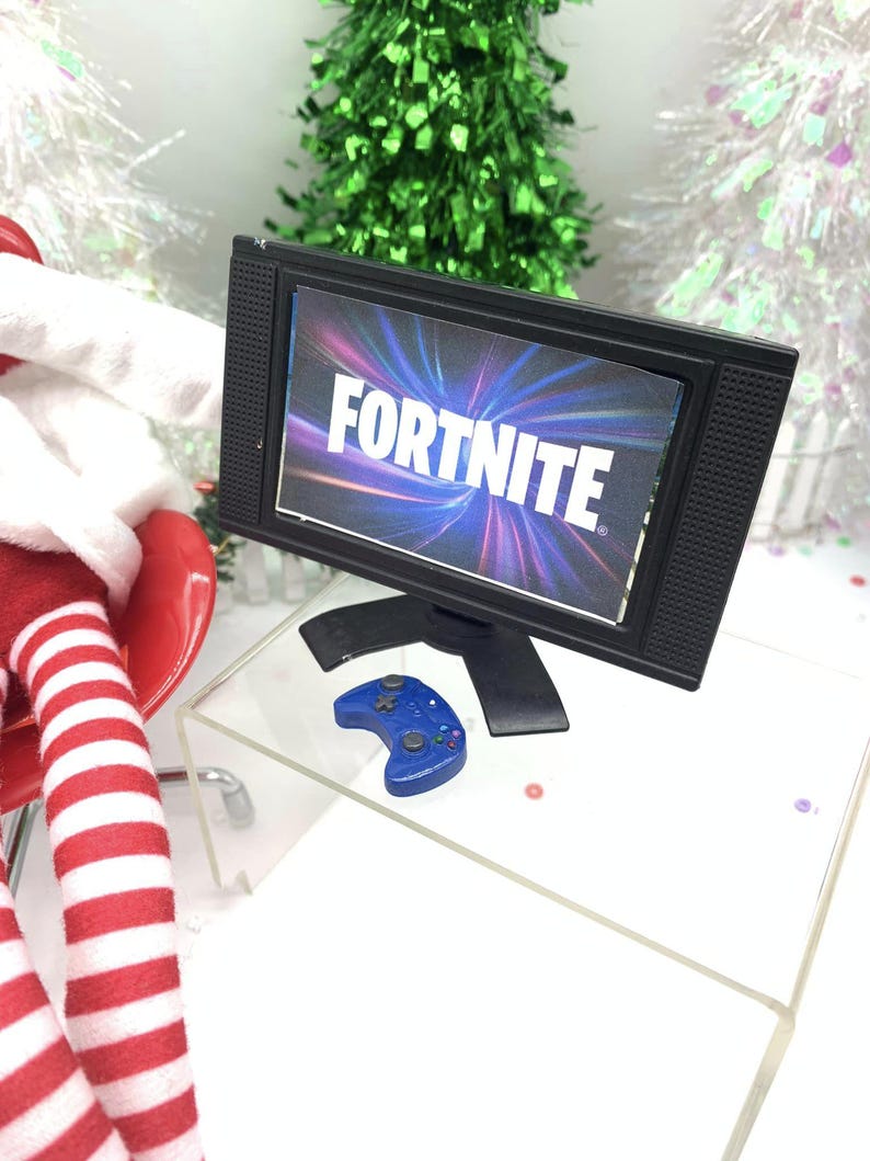 May include: Miniature black television displaying the word "FORTNITE". A blue game controller is positioned below the screen. The TV and controller are on a clear acrylic surface, with Christmas trees in the background.