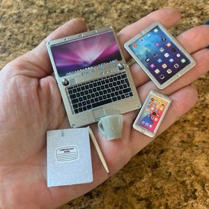 Puede incluir: Un port&aacute;til plateado con el texto "Mini Book" en el teclado, una tableta plateada con una pantalla azul, un tel&eacute;fono plateado con una pantalla blanca, una libreta de composici&oacute;n blanca, una taza blanca y un l&aacute;piz de madera.