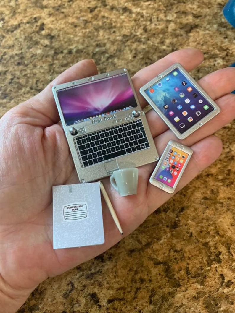 Puede incluir: Un port&aacute;til plateado con el texto "Mini Book" en el teclado, una tableta plateada con una pantalla azul, un tel&eacute;fono plateado con una pantalla blanca, una libreta de composici&oacute;n blanca, una taza blanca y un l&aacute;piz de madera.