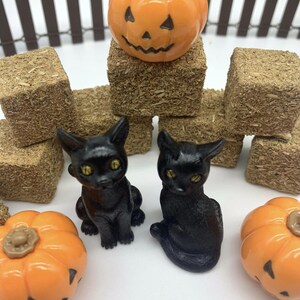 May include: Two black cat figurines with yellow eyes, sitting on a white surface. There are three orange pumpkins with jack-o-lantern faces and hay bales in the background.