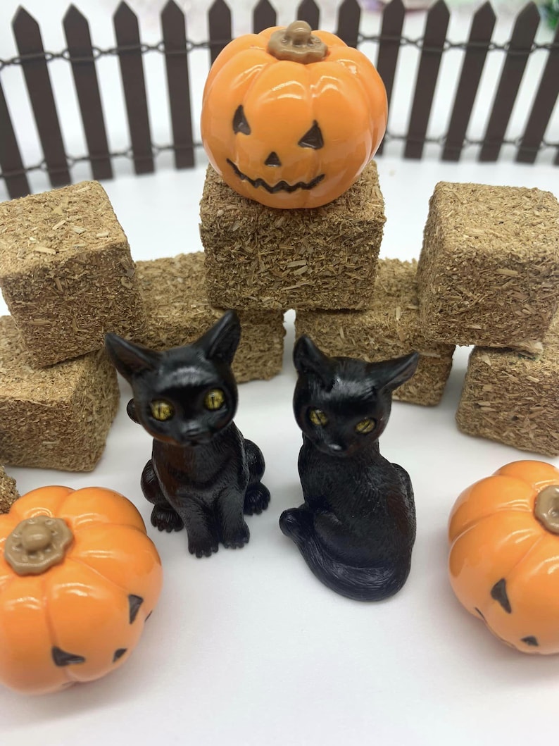 May include: Two black cat figurines with yellow eyes, sitting on a white surface. There are three orange pumpkins with jack-o-lantern faces and hay bales in the background.
