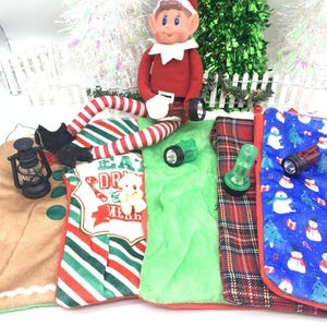 May include: A Christmas elf doll with a red and white outfit holding a flashlight. The elf is sitting on a green plush blanket with four other blankets in the background. The blankets are decorated with Christmas themes, including gingerbread, plaid, and snowmen.