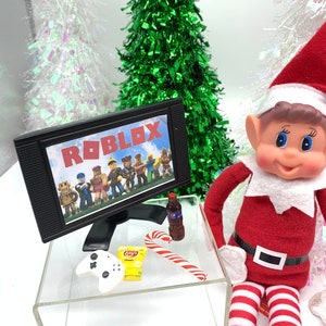 May include: A miniature Christmas scene featuring a toy elf in a red suit and hat. A small TV displays the Roblox logo and characters. Accessories include a candy cane, chips, soda, and a game controller.