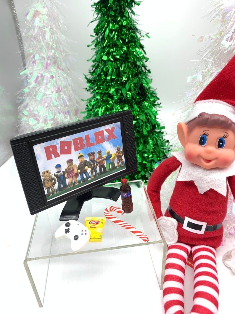 May include: A festive miniature scene with a toy elf in a red outfit. A small TV shows the Roblox logo and characters. The scene includes a candy cane, crisps, a soft drink, and a game controller.