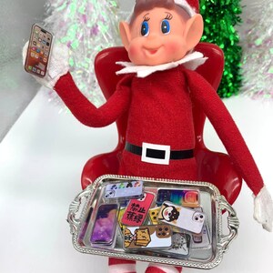 Puede incluir: Un mu&ntilde;eco de duende de Navidad sentado en una silla roja sosteniendo un tel&eacute;fono en miniatura. El duende lleva un atuendo rojo y blanco a rayas y un gorro de Pap&aacute; Noel. El duende sostiene una bandeja plateada con una variedad de tel&eacute;fonos en miniatura.