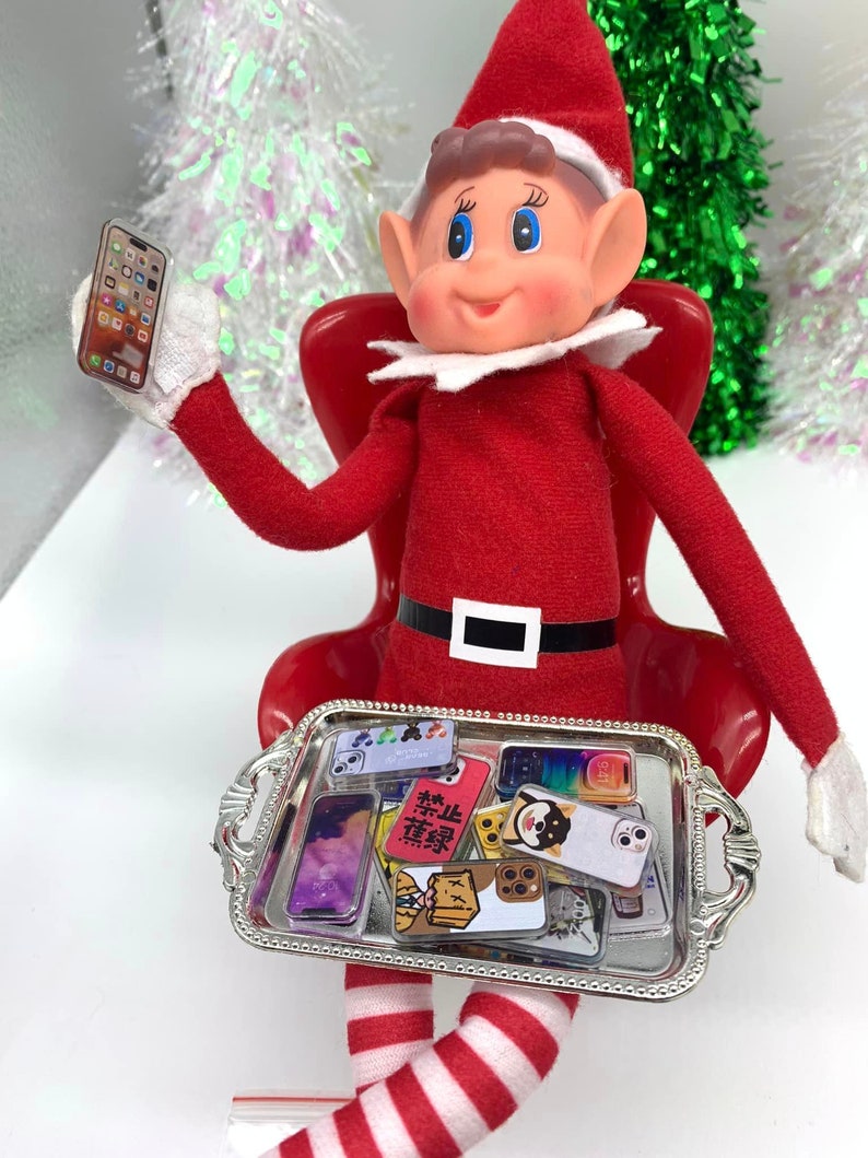 Puede incluir: Un mu&ntilde;eco de duende de Navidad sentado en una silla roja sosteniendo un tel&eacute;fono en miniatura. El duende lleva un atuendo rojo y blanco a rayas y un gorro de Pap&aacute; Noel. El duende sostiene una bandeja plateada con una variedad de tel&eacute;fonos en miniatura.