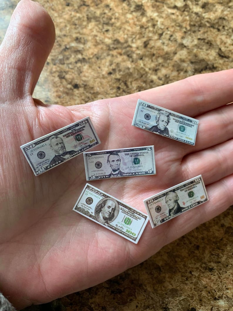 Może przedstawiać: Pięć miniaturowych banknot&oacute;w dolar&oacute;w amerykańskich, w tym banknot 100 dolar&oacute;w, banknot 5 dolar&oacute;w i trzy banknoty 5 dolar&oacute;w. Banknoty są ułożone w dłoni, przy czym banknot 100 dolar&oacute;w znajduje się na dole.