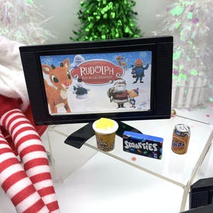 May include: A miniature black television shows a scene from "Rudolph the Red-Nosed Reindeer." Beside it are tiny popcorn, Smarties sweets, and a can of Fanta. The scene is set with miniature Christmas trees.