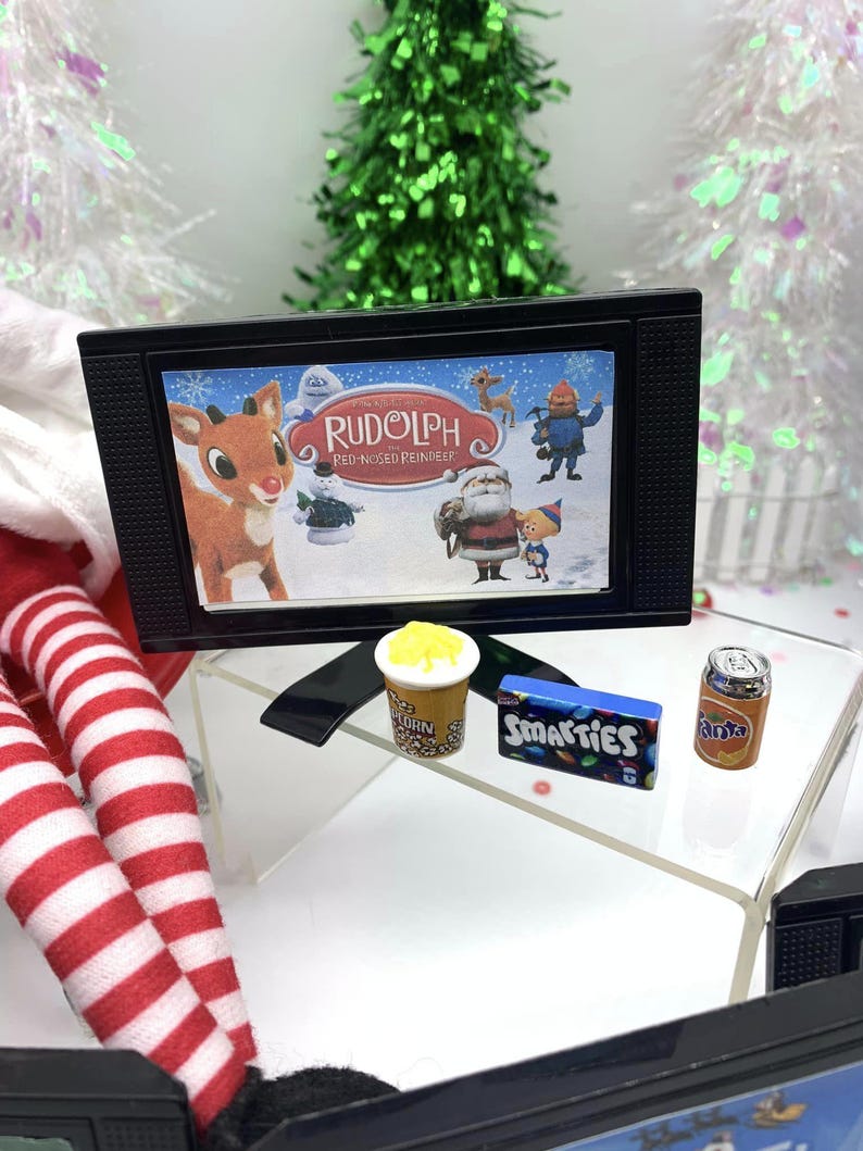 May include: A miniature black television shows a scene from "Rudolph the Red-Nosed Reindeer." Beside it are tiny popcorn, Smarties sweets, and a can of Fanta. The scene is set with miniature Christmas trees.