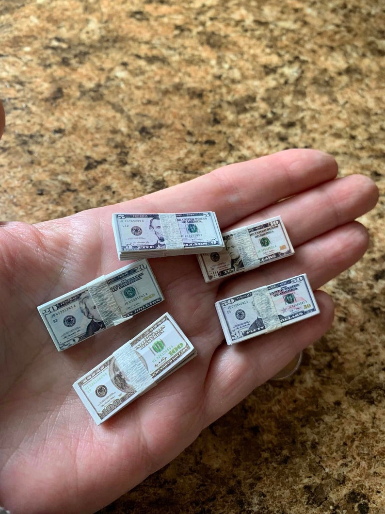 Może przedstawiać: Pięć stos&oacute;w miniaturowych banknot&oacute;w dolar&oacute;w amerykańskich, w tym nominały 5, 10, 20, 50 i 100 dolar&oacute;w, jest wyświetlanych na dłoni. Banknoty są nadrukowane wizerunkiem Abrahama Lincolna, Alexandra Hamiltona, Andrew Jacksona, Ulyssesa S. Granta i Benjamina Franklina.
