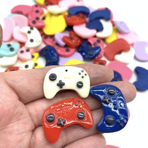 May include: Three miniature plastic game controllers in white, red, and blue. The controllers have black buttons and colorful accents. The controllers are on a hand.