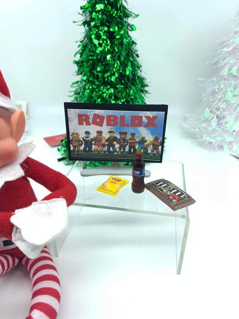 May include: A miniature scene with a television showing "ROBLOX" characters. A bag of crisps, a bottle of fizzy drink, and a chocolate bar are on a clear table. A green tinsel Christmas tree is in the background.