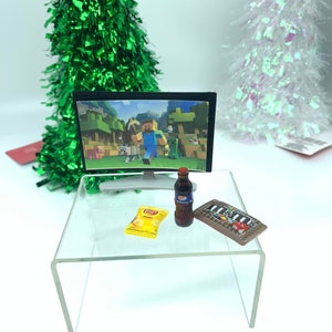 May include: Miniature scene featuring a TV showing Minecraft, a Pepsi bottle, a bag of Lay's crisps, and a chocolate bar on a clear acrylic table. Two decorative Christmas trees are in the background.