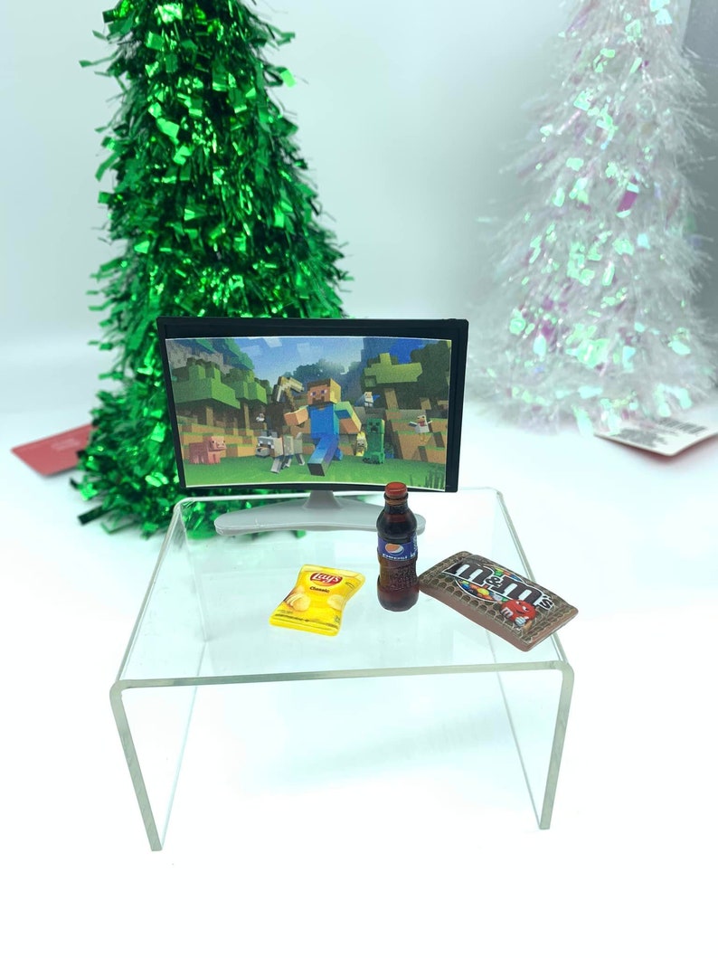 May include: Miniature scene featuring a TV showing Minecraft, a Pepsi bottle, a bag of Lay's crisps, and a chocolate bar on a clear acrylic table. Two decorative Christmas trees are in the background.