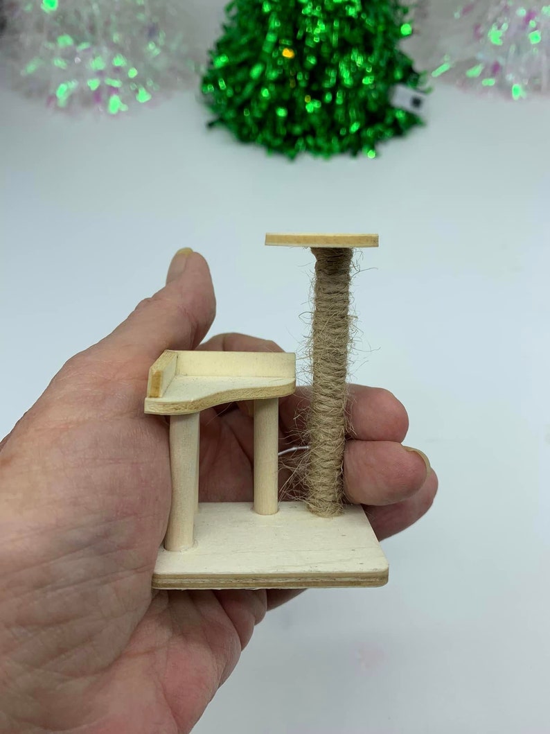 May include: A miniature wooden cat tree with a sisal-covered scratching post and two platforms. The cat tree is small enough to fit in the palm of a hand.