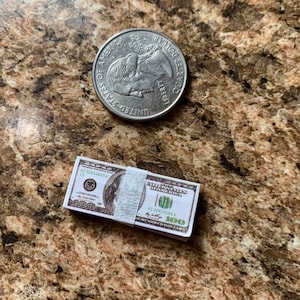 Może przedstawiać: Stos dziesięciu podrobionych banknot&oacute;w 100-dolarowych z białą gumką. Banknoty są nadrukowane tekstem "THE UNITED STATES OF AMERICA" i "ONE HUNDRED DOLLARS". Na zdjęciu znajduje się r&oacute;wnież srebrna moneta 25-centowa.