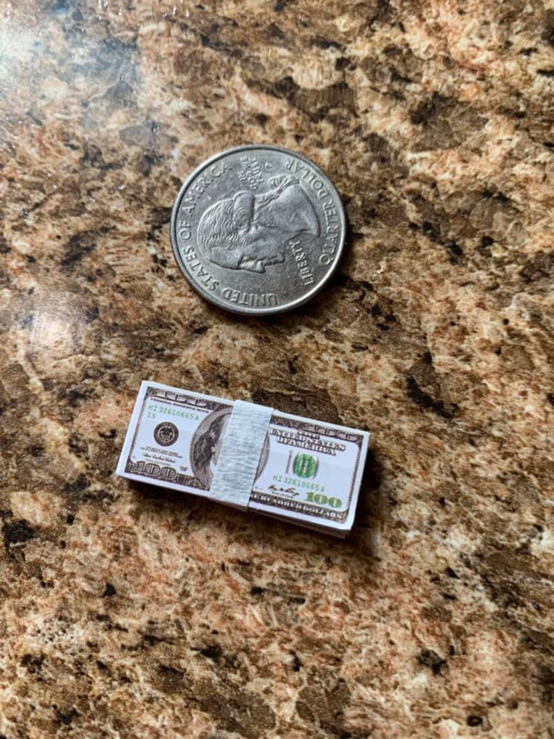 Może przedstawiać: Stos dziesięciu podrobionych banknot&oacute;w 100-dolarowych z białą gumką. Banknoty są nadrukowane tekstem "THE UNITED STATES OF AMERICA" i "ONE HUNDRED DOLLARS". Na zdjęciu znajduje się r&oacute;wnież srebrna moneta 25-centowa.